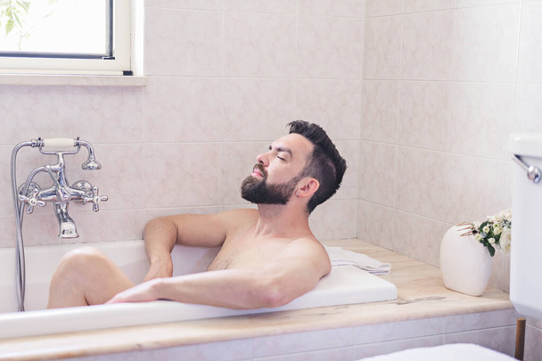 Бородатый мужчина наслаждается и расслабляется во время купания в бежевой винтажной ванной. Концепция ухода за кожей и кожей, мужское здоровье - Фото, изображение
