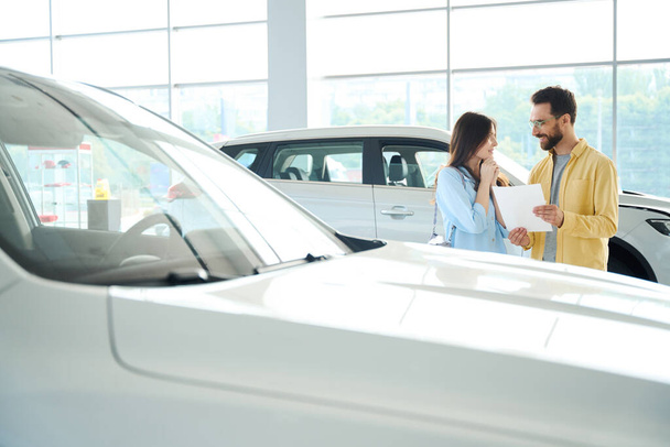 Diligente hombre en gafas mostrando papel a su hermosa novia en el centro de coches - Foto, Imagen