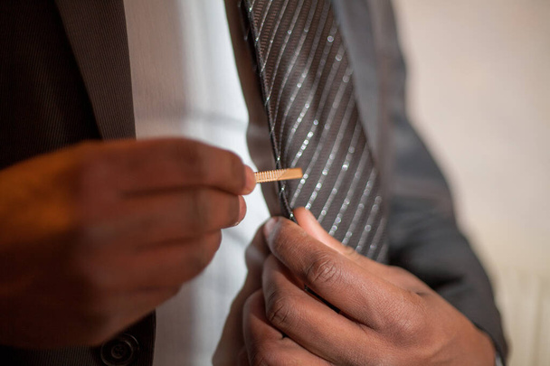 Nesta imagem, um close-up captura o momento preciso em que um homem segura um grampo de gravata em sua gravata listrada. A ação está focada nas mãos, habilmente aplicando o acessório, que é funcional e - Foto, Imagem
