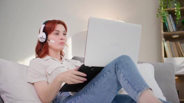 Nuori nainen toimii internet-kuulokkeilla ja kannettavalla tietokoneella, kommunikoi videoviestinnän kautta istuessaan sängyssä makuuhuoneessa - Materiaali, video