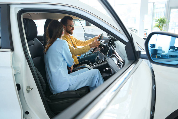 Handsome man in glasses and pretty female in stylish clothes sitting in car interior in car showroom - Photo, Image