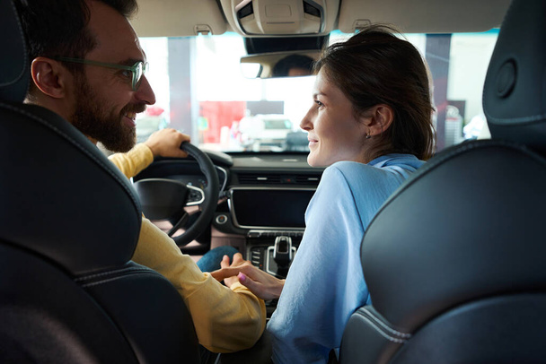 Uśmiechnięty mężczyzna w okularach i biała kobieta patrząc na siebie podczas siedzenia w samochodzie wnętrza w salonie samochodowym - Zdjęcie, obraz