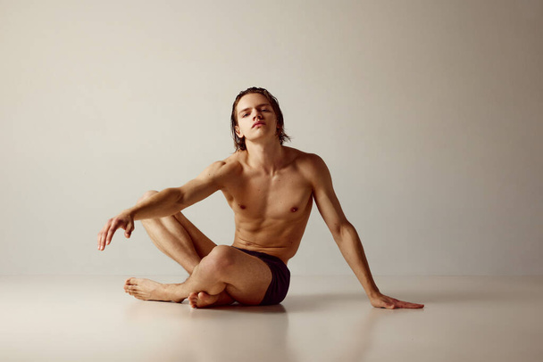 Handsome young guy with sportive, muscular body sitting on floor, posing shirtless in underwear against grey studio background. Concept of mens beauty, health, body care, sportive lifestyle - Zdjęcie, obraz