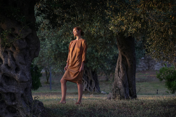 Beautiful young woman in goolden colored silk dress standing next to the old olive tree - Photo, Image