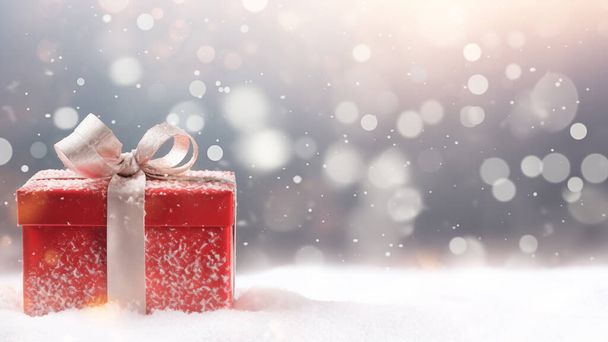 Een rode geschenkdoos met een zilveren lint, genesteld in sneeuw met een bokeh licht decor - Foto, afbeelding