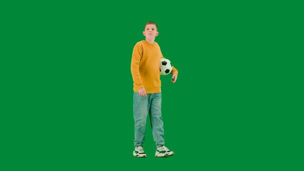 School kids and leisure time creative concept. Portrait of kid boy on chroma key green screen. Schoolboy in jeans walking holding football ball and looking around. Full body profile shot. - Foto, Bild