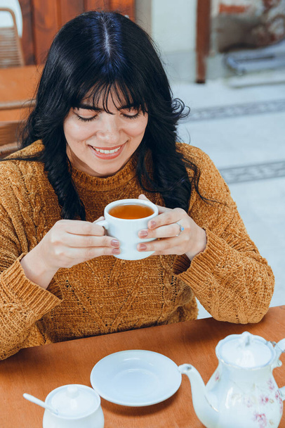 Beyaz porselen bardakla oturmuş çay içen güzel Latin genç bir kadının manzarası. Gülümsüyor ve günün tadını çıkarıyor.. - Fotoğraf, Görsel