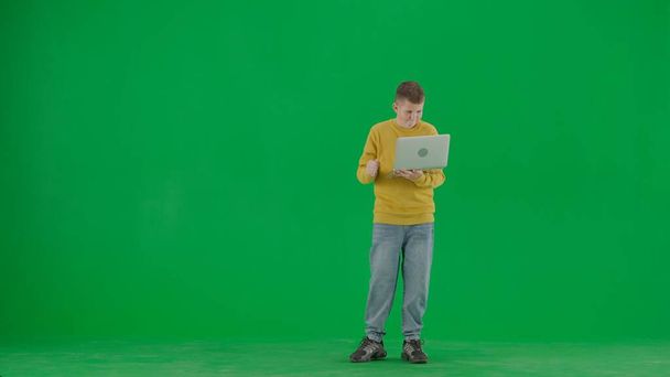 School kids and leisure time creative concept. Portrait of kid boy on chroma key green screen. Schoolboy in jeans holding laptop and looking with winning face expression. Front body front shot. - Photo, Image