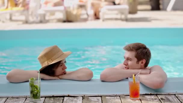 プールで会話をする若い恋人のカップル. 男性と女性は青い水のプールサイドでアペロールを浴びて飲んでいます. 夏の晴れた日についての家族休暇の会話. ハイ ハイ - 映像、動画