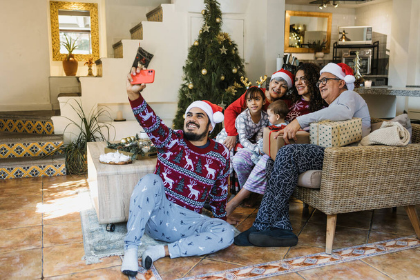 Familia latina tomando retrato selfie con teléfono móvil durante la Navidad en casa en México América Latina. gente hispana abuelos hija, madre y padre con sombrero de santa - Foto, Imagen
