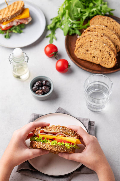Handen met een clubsandwich met spek, kaas, tomaat en sla op een lichte achtergrond met olijven, kruiden en een glas water. Gezond snackconcept voor ontbijt of lunch. Bovenaanzicht. - Foto, afbeelding
