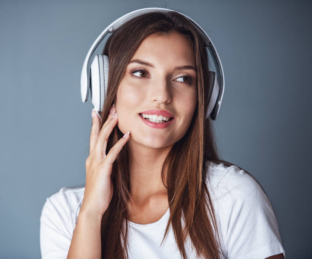 Attrayant jeune femme brune dans les écouteurs écoute de la musique et souriant, sur fond gris - Photo, image