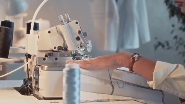 Plan latéral recadré de couturière méconnaissable travaillant avec une machine à coudre industrielle faisant des coutures noires sur du textile blanc pendant la journée de travail à l'usine de vêtements - Séquence, vidéo