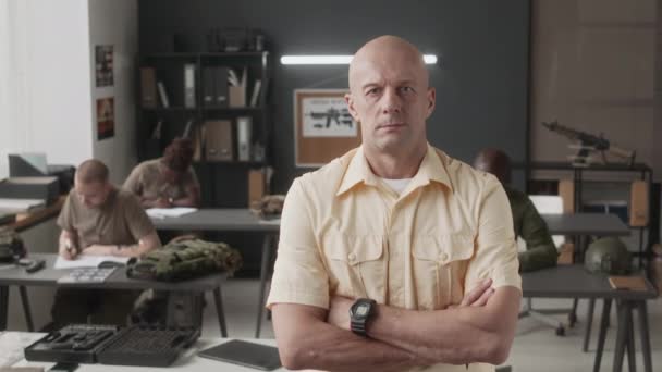 Portrait moyen d'un instructeur de service militaire caucasien chauve posant les bras croisés en classe et regardant une caméra avec des cadets en arrière-plan - Séquence, vidéo