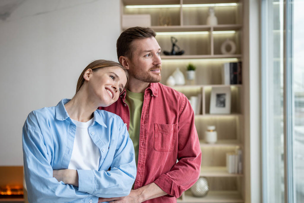 Amiable cheerful happy woman and tender caring attentive man standing in silence in own flat, looking at window, dreaming, thinking on relations, love, family, togetherness, future plans, children  - Photo, Image