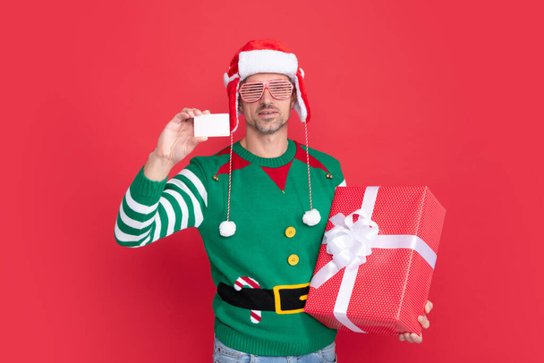happy xmas guy with present box on red background. happy new year. merry christmas gift. man holding debit card for shopping. man in elf costume and santa hat with glasses. - Photo, Image