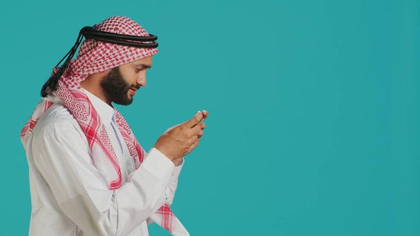 Radosny muzułmanin bawiący się z gry online na swoim smartfonie, w tradycyjnym islamskim stroju. W studiu, arabski dorosły korzystających e wyzwanie sportowe z telefonu komórkowego urządzenia. - Zdjęcie, obraz