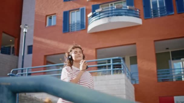 Słuchawki nastolatek tańczący schody uliczne w letni dzień. Beztroska kręcona dziewczyna słuchająca muzyki w słuchawkach poruszających się w taktowej piosence na świeżym powietrzu. Modna hipsterka trzymająca magnetofon retro na schodach - Materiał filmowy, wideo