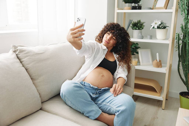 Zwangere vrouw blogger zit op de bank thuis en neemt foto 's van zichzelf op de telefoon, selfie en video gesprek, overleg met de arts online, zwangerschap management. Hoge kwaliteit foto - Foto, afbeelding