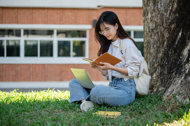 Una hermosa joven asiática estudiante universitaria en ropa casual está haciendo deberes, investigando información en un libro y usando su computadora portátil bajo el árbol en un parque del campus. - Foto, Imagen