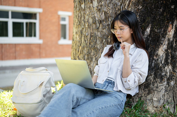 Eine positive und nachdenkliche asiatische Studentin benutzt ihren Laptop unter dem Baum in einem Campus-Park und macht online Hausaufgaben. - Foto, Bild
