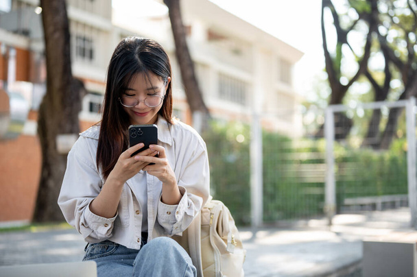 Положительная и обаятельная молодая азиатка в повседневной одежде болтает со своими друзьями на смартфоне, отдыхая на скамейке в парке. Люди и технологии - Фото, изображение