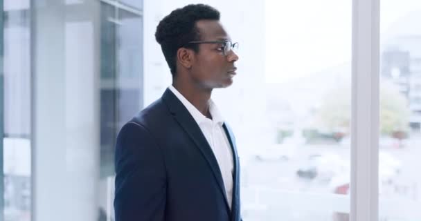 Gezicht, zaken en een serieuze zwarte man voor een raam in zijn kantoor voor professioneel werk of carrière. Portret, bedrijf en bril met een zelfverzekerde jonge werknemer in een pak op zijn werkplek in de dag. - Video