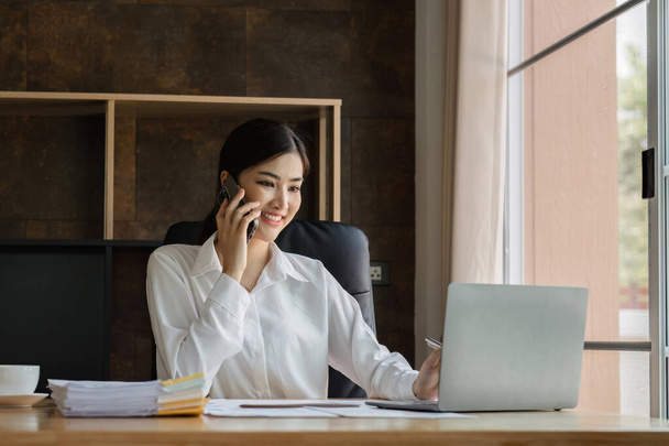 Lachende Aziatische zakenvrouw gebruikt laptop om te werken aan financiële transacties. Planning van het bureau op kantoor. - Foto, afbeelding