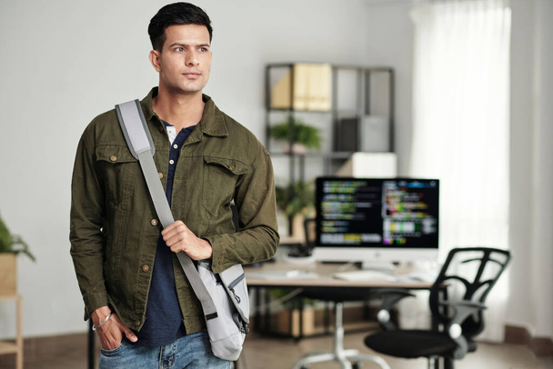 Porträt eines jungen, ehrgeizigen Software-Entwicklers mit Kreuztasche, der im Büro steht und wegschaut - Foto, Bild