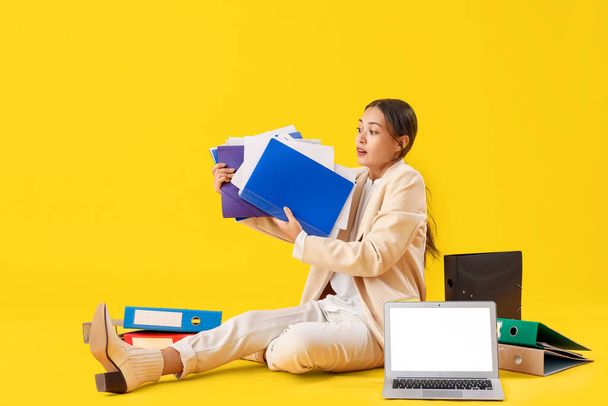 Νεαρή Ασιάτισσα επιχειρηματίας με φακέλους εγγράφων και φορητό υπολογιστή σε κίτρινο φόντο - Φωτογραφία, εικόνα