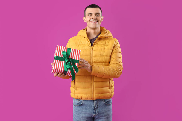 Jongeman in warm kogelvrij vest met kerstcadeau op paarse achtergrond - Foto, afbeelding