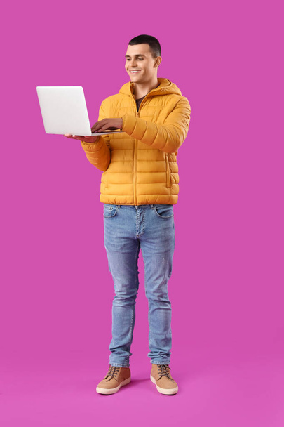 Young man in warm puffer jacket using laptop on purple background - Photo, Image