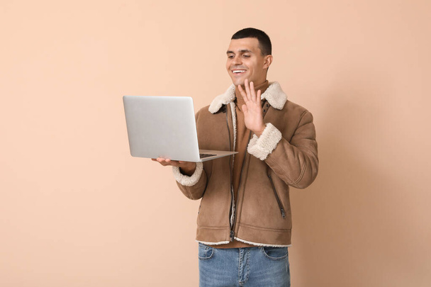 Молодой человек в бараньей куртке с помощью ноутбука на бежевом фоне - Фото, изображение