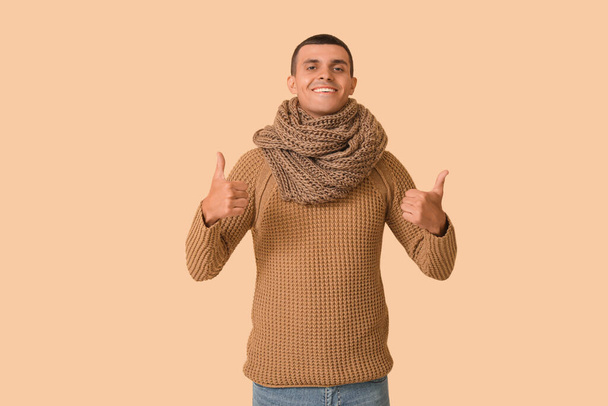 Hombre joven en ropa de invierno mostrando pulgares hacia arriba sobre fondo beige - Foto, imagen