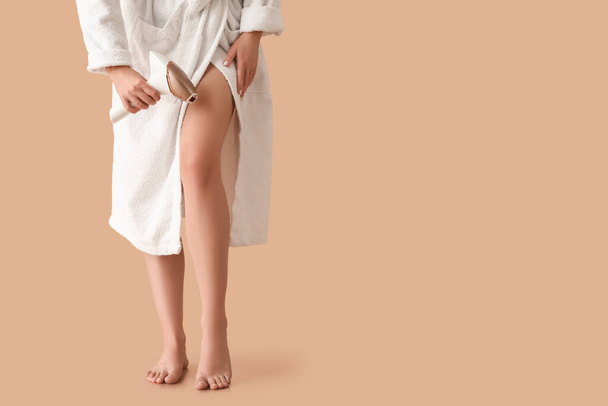 Piękna młoda kobieta w szlafroku depiluje nogi fotodepilatorem na brązowym tle - Zdjęcie, obraz