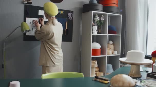 Mittlere Gesamtaufnahme einer jungen kaukasischen Hutmacherin, die in ihrer Werkstatt herumläuft, Kerzen anzündet und ihre neuen Entwürfe betrachtet - Filmmaterial, Video
