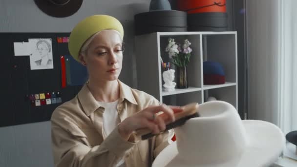 Mittlere Aufnahme einer kaukasischen Designerin, die ihren neuen weißen Hut ausbürstet - Filmmaterial, Video