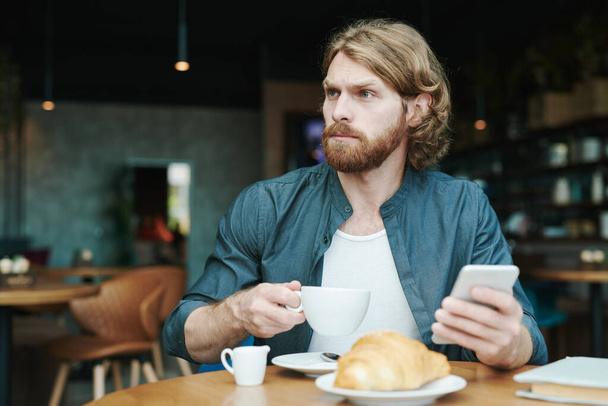 Serious handsome guy with beard sitting at table in cafe and drinking coffee while reading mail on smartphone - Zdjęcie, obraz