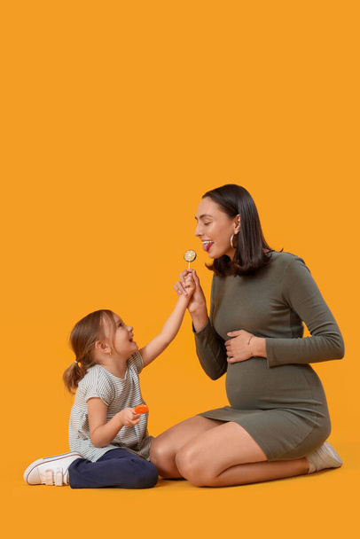 Piękna ciężarna matka i jej słodka córeczka ze słodkimi lizakami siedzącymi na żółtym tle - Zdjęcie, obraz