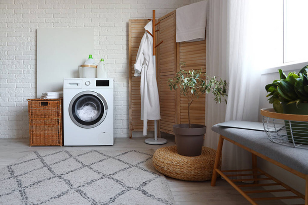 Interior da moderna lavanderia com máquina de lavar roupa, roupão de banho em rack e cesta de vime - Foto, Imagem