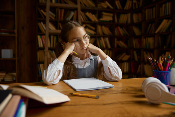 Portrait of pensive girl student doing homework or studying at desk in home library. Homeschooling concept - Фото, зображення