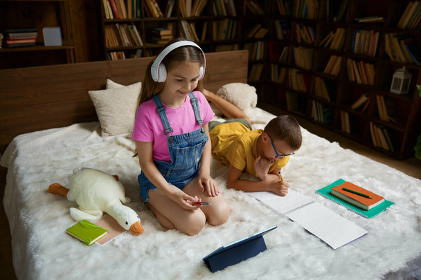 Happy schoolkid studying and preparing homework together at home. Little sister listening teacher using headset and digital tablet device while her brother writing task in copybook - Photo, Image
