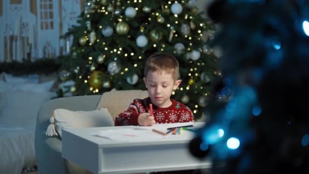 Ένα λευκό αγόρι γράφει ένα γράμμα στον Άγιο Βασίλη κοντά στο χριστουγεννιάτικο δέντρο στο σπίτι. Ένα χαρούμενο παιδί ετοιμάζεται για διακοπές και δώρα στην ατμόσφαιρα ενός παραμυθιού. Υψηλής ποιότητας 4k πλάνα - Πλάνα, βίντεο