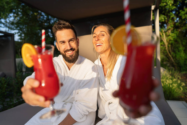 vrolijk paar in witte spa gewaden genietend van rode cocktails in een wellness resort - Foto, afbeelding