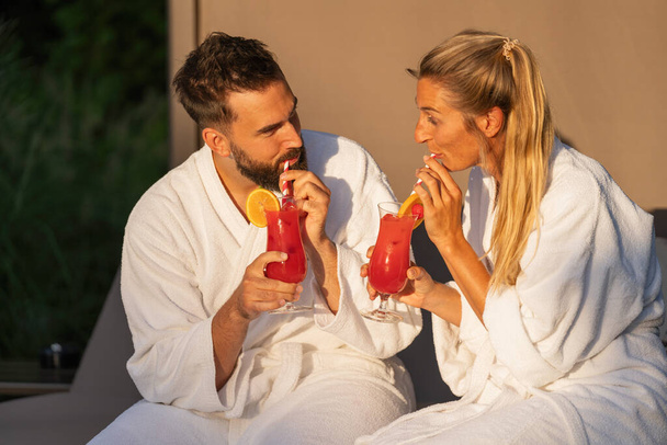 Uomo e donna in accappatoi bianchi sorseggiando cocktail in luce ora d'oro al resort spa hotel - Foto, immagini