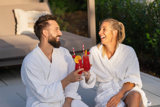 radosna para w białych szlafrokach brzęcząca szklankami z czerwonymi napojami, na świeżym powietrzu o złotej godzinie w hotelu - Zdjęcie, obraz