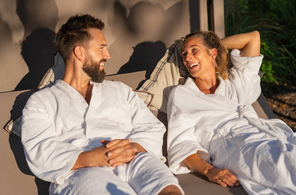 Pareja con túnicas blancas descansando y disfrutando de una risa en una tumbona a la luz del sol en un hotel spa  - Foto, imagen