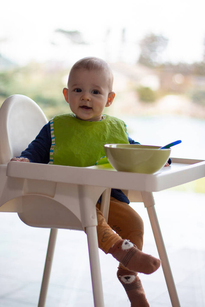 Pequena criança comendo em cadeira alta, menino feliz vista de corpo inteiro, aprendendo a comer e tem comida no rosto - Foto, Imagem