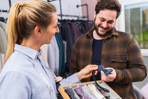 Χαμογελώντας ράφτης δείχνει ένα μπλε παπιγιόν σε ένα ευτυχισμένο αρσενικό πελάτη, κοστούμια στο παρασκήνιο - Φωτογραφία, εικόνα