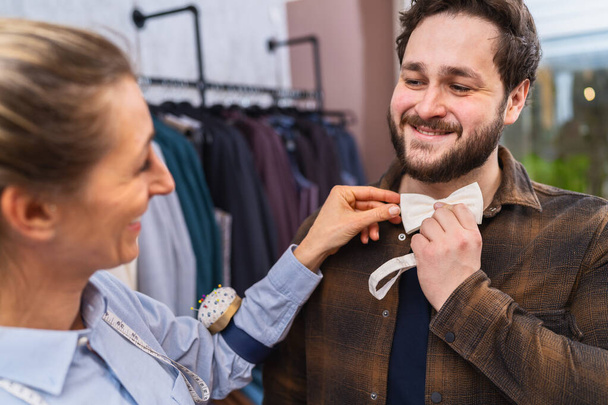 Tailor προσαρμογή ένα παπιγιόν για ένα χαμογελαστό αρσενικό πελάτη, ράφια ρούχα σε ένα κατάστημα γάμου - Φωτογραφία, εικόνα